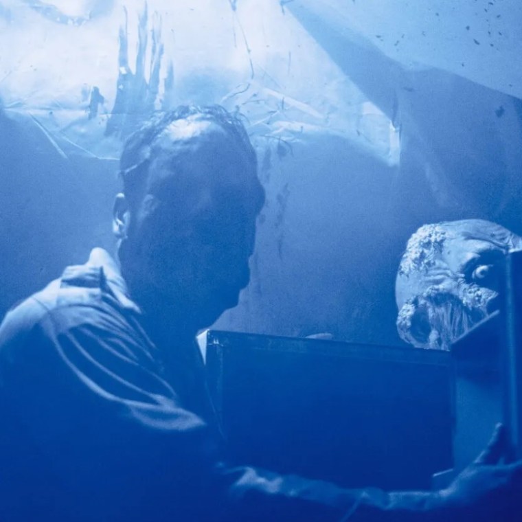 Man in blue light with zombie mask peering over shoulder in eerie setting.
