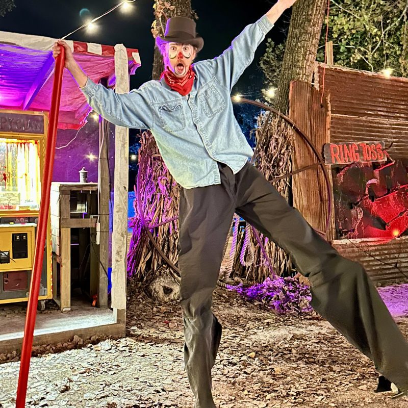 Person on stilts in costume with top hat, outdoors at night with colorful lights.