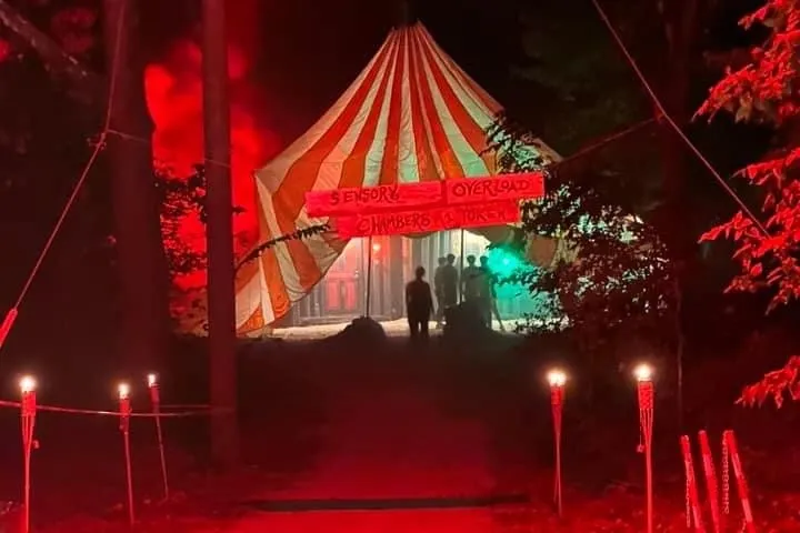 Red-lit path leads to a striped circus tent entrance in a dark forest.