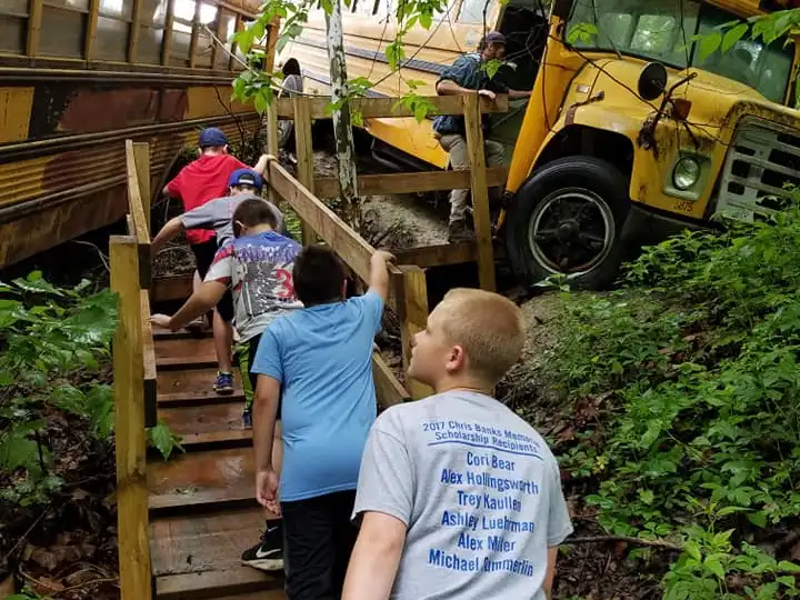 Children climbing wooden steps beside old school buses in a forest setting.
