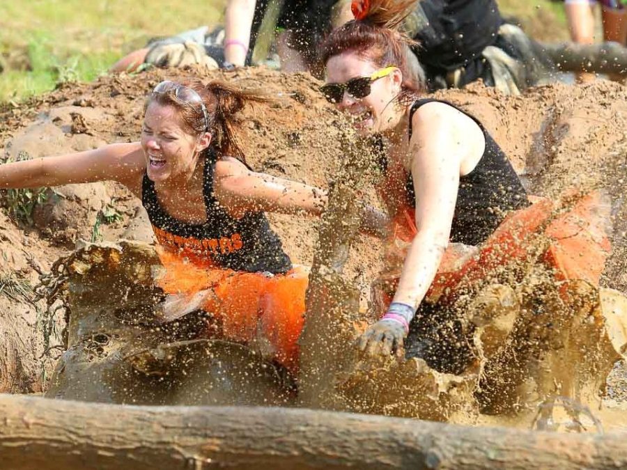 Women in black tank tops and orange skirts splash through muddy water in an outdoor obstacle course.
