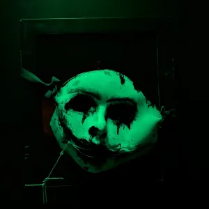 Green-lit mask with black paint drips, mounted on a dark wall.