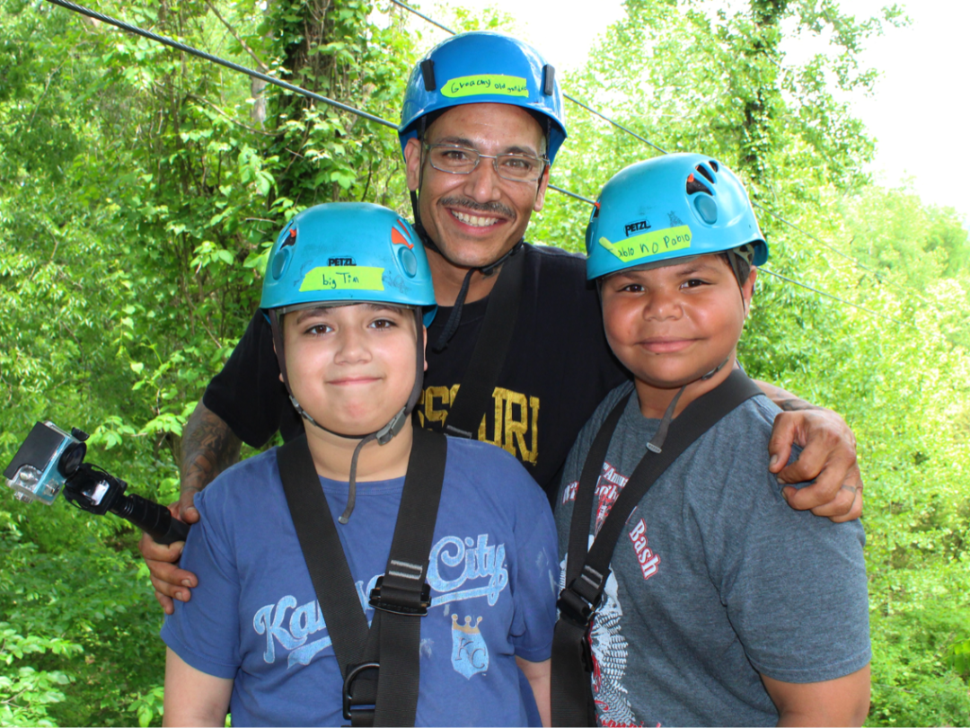 Three people wearing helmets and harnesses smiling in a forest setting.