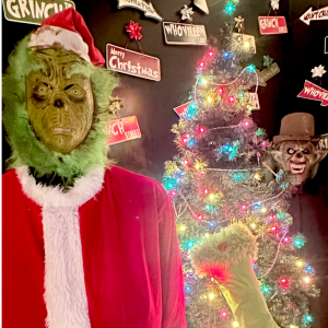 Two people in Christmas costumes beside a decorated tree and festive signs.