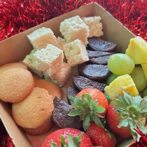 Box of strawberries, grapes, pineapple, cookies, and rice crisp squares with dip, surrounded by red tinsel.
