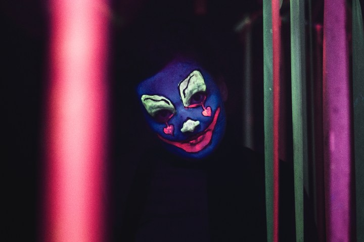A figure in a dark room with a glowing clown face, vivid colors, and striped pants.