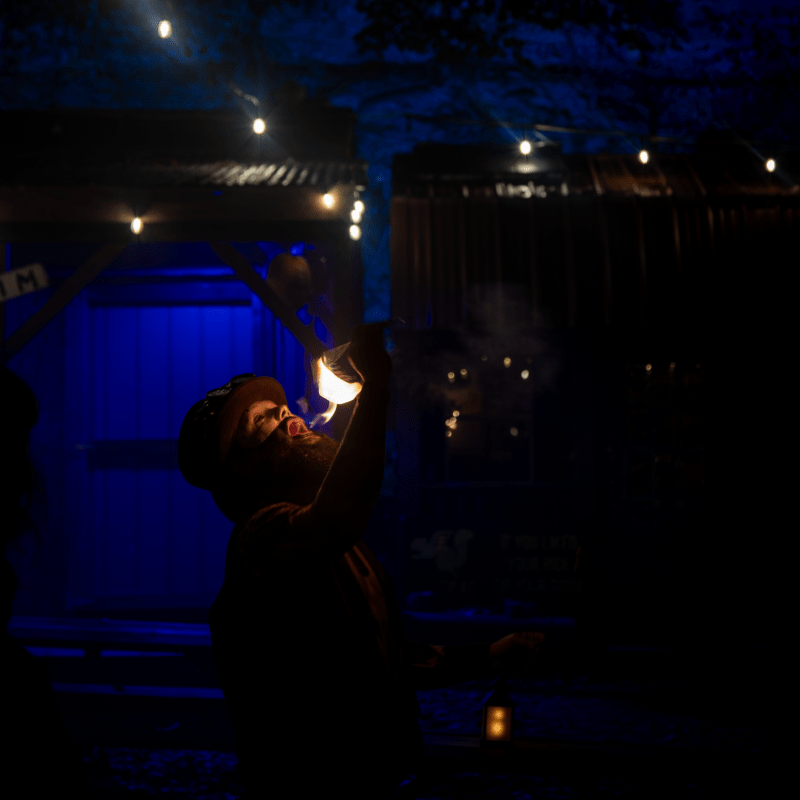 Performer breathes fire under string lights in dark setting.