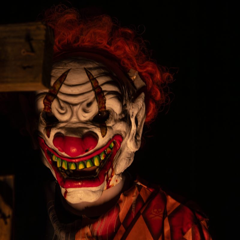 Creepy clown with orange hair, sharp teeth, and horned mask in dark setting, wearing diamond pattern outfit.