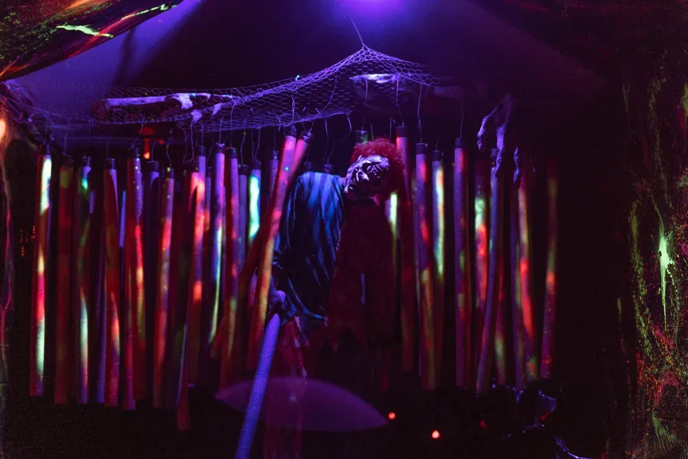 An Exiled clown waits among hanging neon tubes and cobwebs